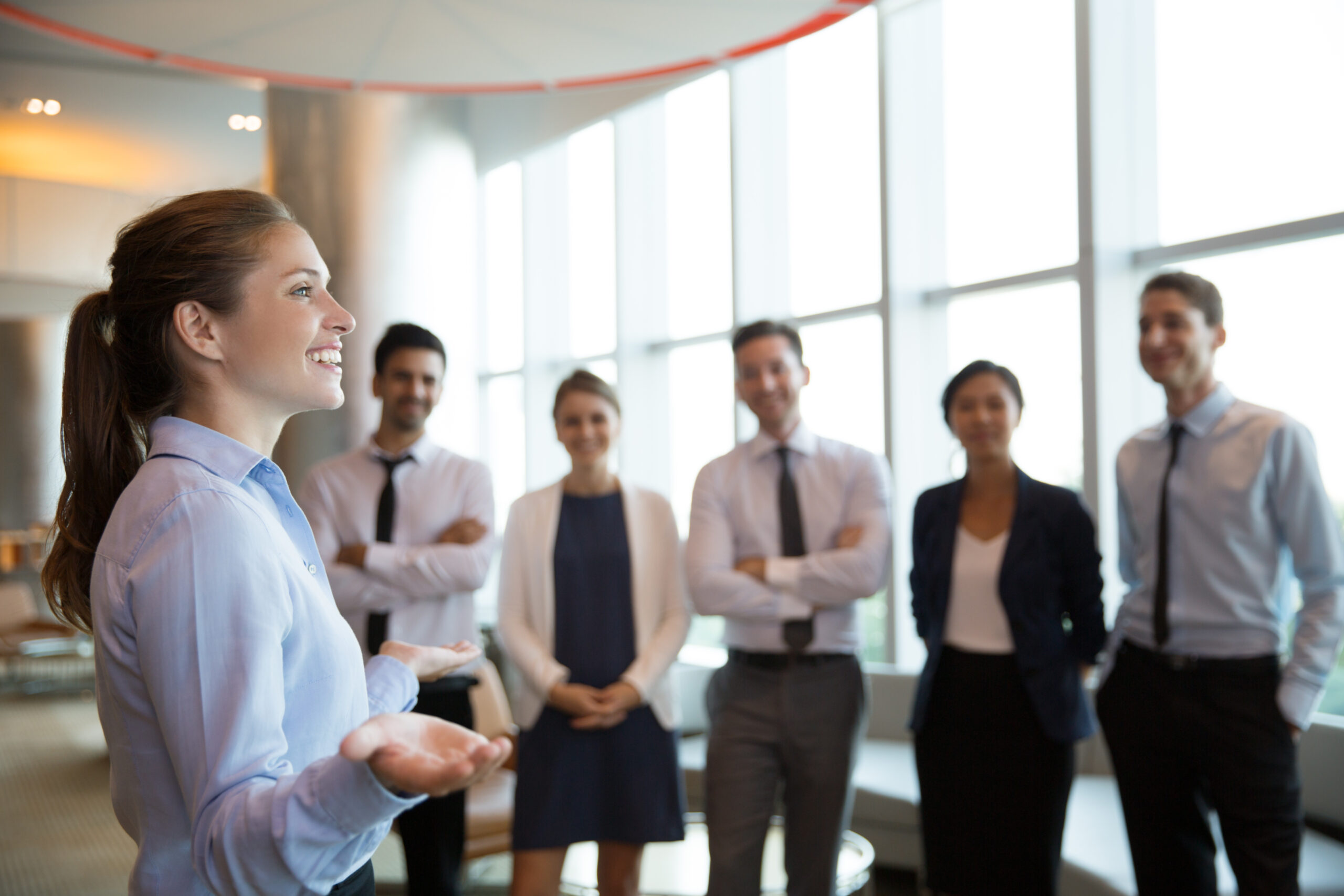 Portrait of successful female executive manager with open hands gesture standing in front of her team in office hall and announce good news
